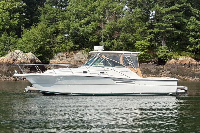 The Image of 2000 Pursuit 3400 Express boat docked near rocky shoreline and lush trees. - 0
