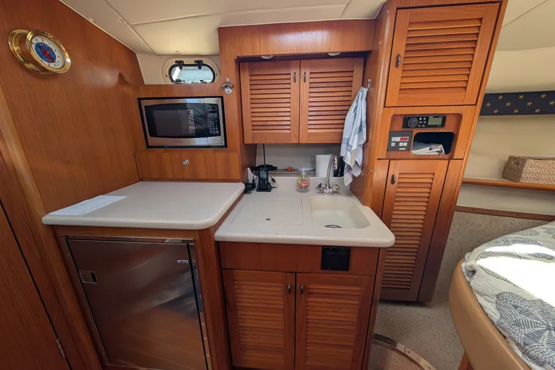 Slide: The Image of Compact kitchen area in a 2000 Pursuit 3400 Express boat, featuring wood cabinetry and modern appliances. - 9
