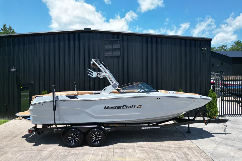 The Image of 2026 MasterCraft XT24 boat on trailer, parked outside a black building under a blue sky. - 1