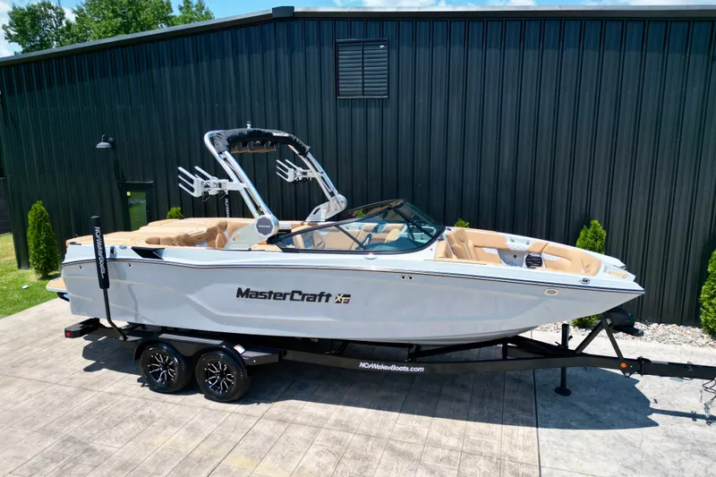 Slide: The Image of 2026 MasterCraft XT24 boat on trailer, parked outdoors near a black building. - 2