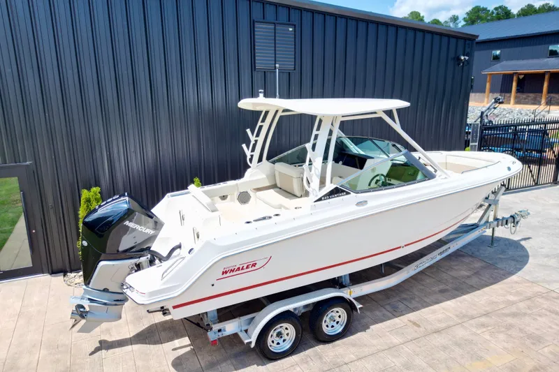 Slide: The Image of 2025 Boston Whaler 240 Vantage boat on trailer, parked near a modern building. - 2