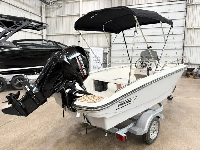 The Image of A 2027 Boston Whaler 130 Sport in a warehouse. It features a black canopy and a Mercury engine. - 0