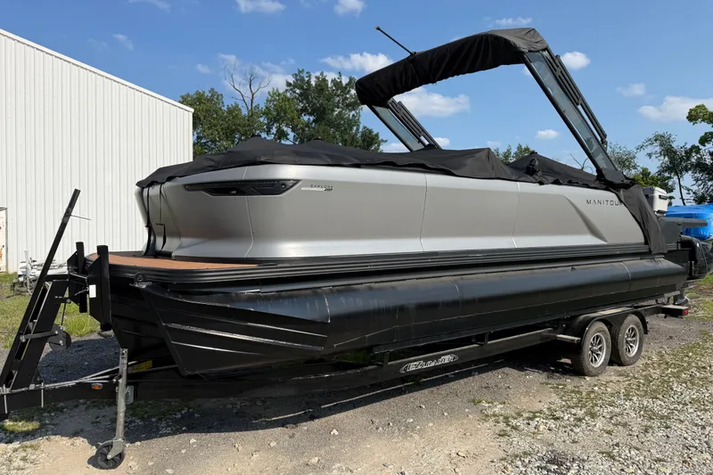 Slide: The Image of 2024 Manitou Explore Dual Engine boat on trailer, parked outdoors under blue sky. - 1