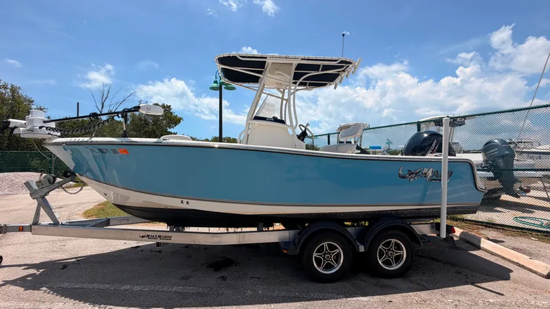 The Image of 2024 Mako 214CC on a trailer, featuring a blue hull and T-top, parked outdoors. - 0