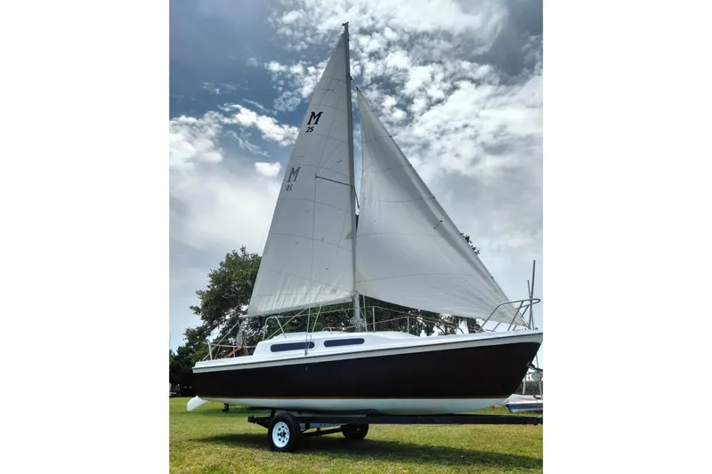 Slide: The Image of 1980 MacGregor 25 sailboat on trailer, white sails, parked on grassy area under cloudy sky. - 1