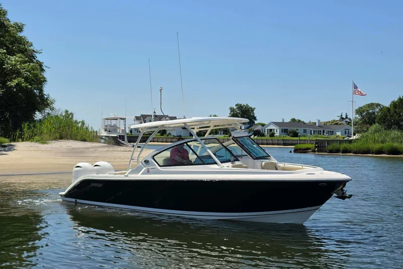 The Image of 2021 Pursuit DC 266 Dual Console boat cruising near a sandy shoreline and residential area. - 0