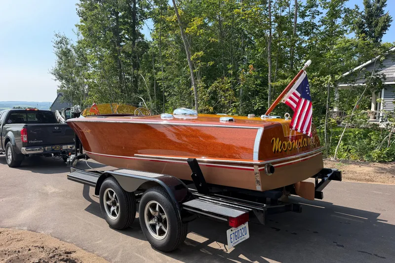 Slide: The Image of 1953 Chris Craft Riviera boat on trailer, American flag, parked near trees. - 3