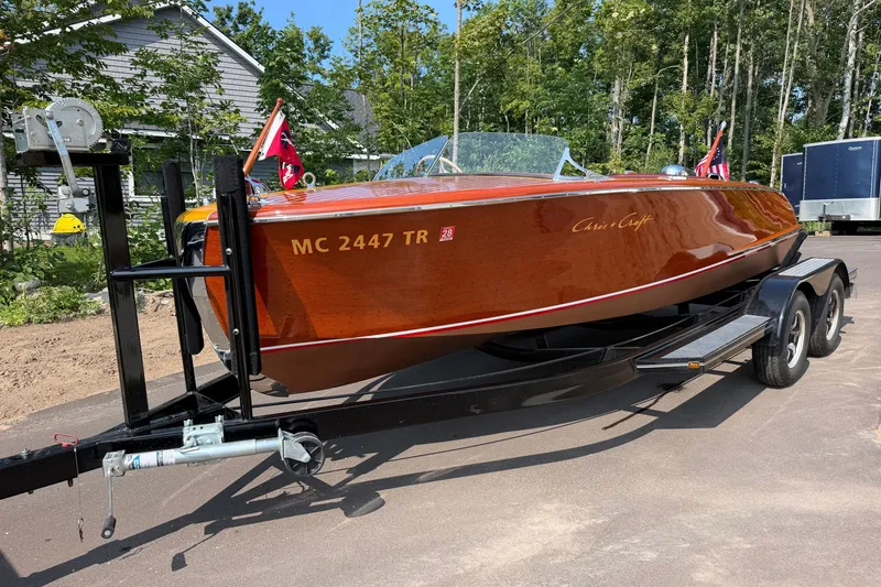 The Image of 1953 Chris Craft Riviera boat on trailer, surrounded by trees and a house. - 1