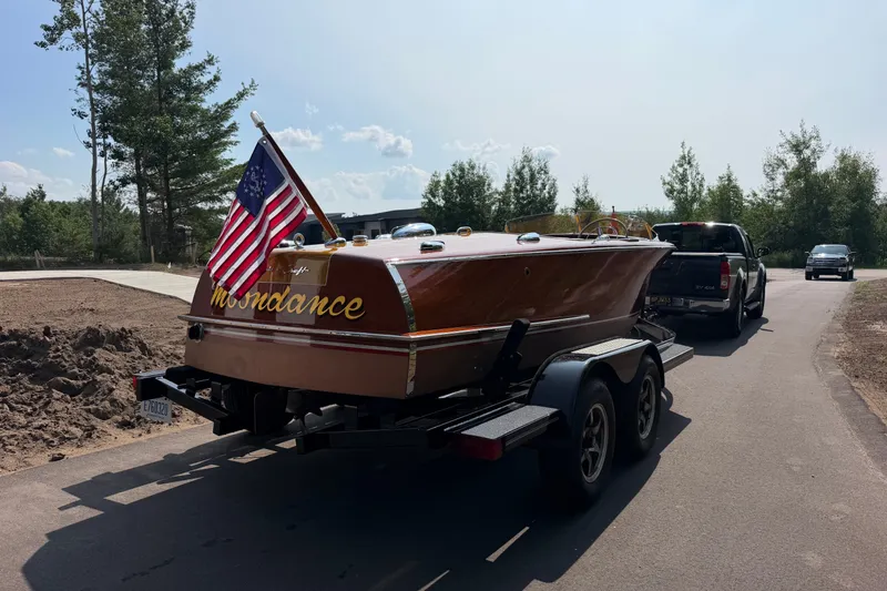 Slide: The Image of 1953 Chris Craft Riviera boat on trailer, displaying American flag, parked on a sunny road. - 5