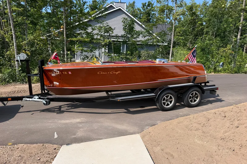 Slide: The Image of 1953 Chris Craft Riviera boat on trailer, parked near a house with trees in the background. - 2