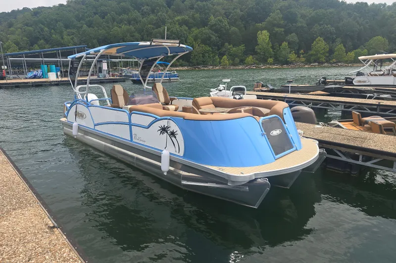 The Image of Aloha 230 Club 2027 pontoon boat docked on a serene lake with lush greenery. - 1