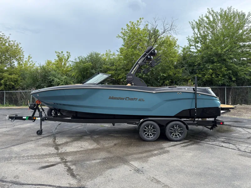 The Image of A 2027 MasterCraft NXT23 boat on a trailer, surrounded by trees. - 0