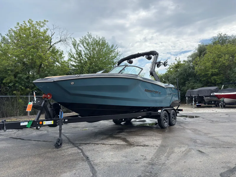 Slide: The Image of A modern 2027 MasterCraft NXT23 on a trailer, surrounded by trees and a cloudy sky. - 1