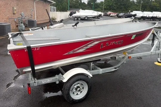 Slide: The Image of Red 2025 Lund WC-12 boat on trailer, parked outdoors on wet pavement. - 13