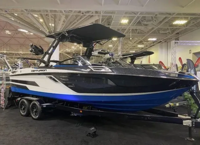 Slide: The Image of 2023 Centurion Ri245: A sleek, modern boat with a blue hull and black canopy, displayed indoors. - 2