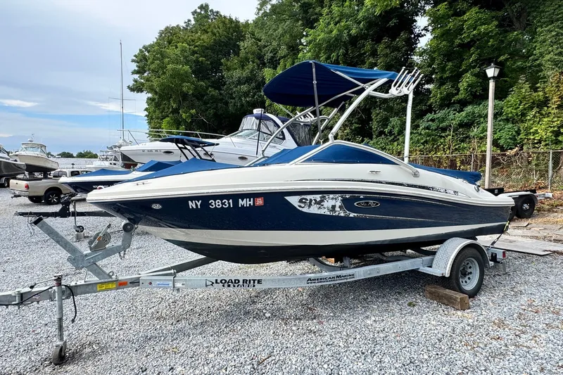 The Image of 2010 Sea Ray 185 Sport boat on trailer, parked in a gravel lot with trees in background. - 0