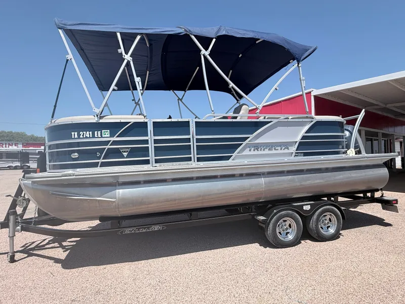 Slide: The Image of A 2018 Trifecta pontoon boat with a blue canopy on a trailer, parked on a gravel lot. - 2