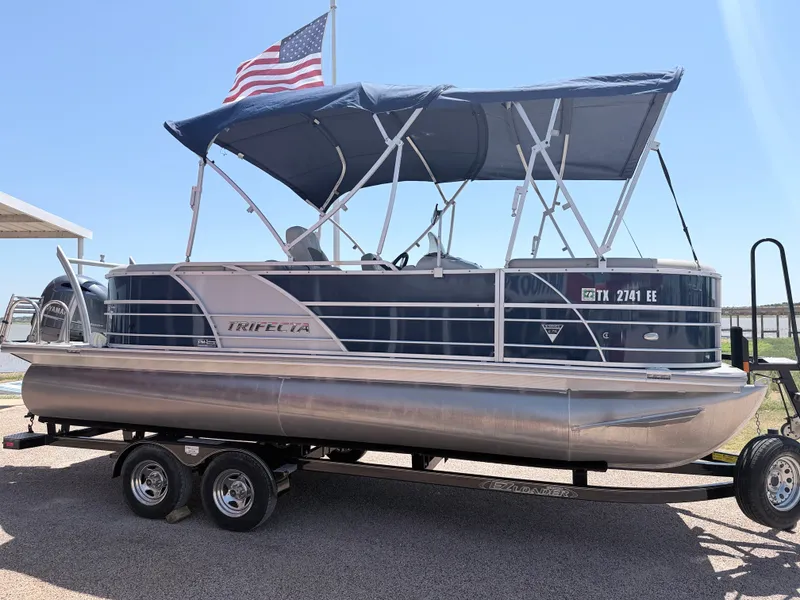 The Image of A 2018 Trifecta pontoon boat with a blue canopy and American flag, on a trailer. - 1