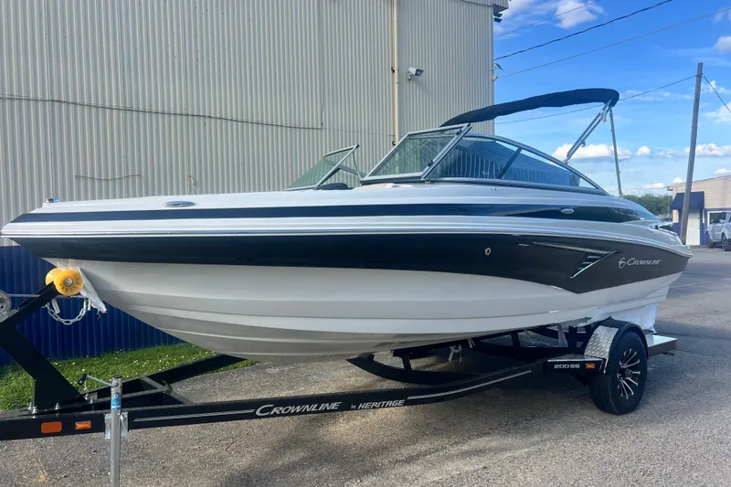 The Image of 2027 Crownline 200 SS boat on trailer, parked outdoors near industrial building. - 0