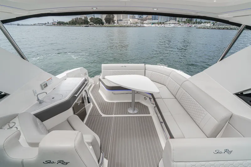 Slide: The Image of The image shows the cockpit of a 2025 Sea Ray 320 Sundancer DA Coupe. It features an elegant seating area with a table and a small kitchenette. Water and a cityscape are visible in the background. - 4