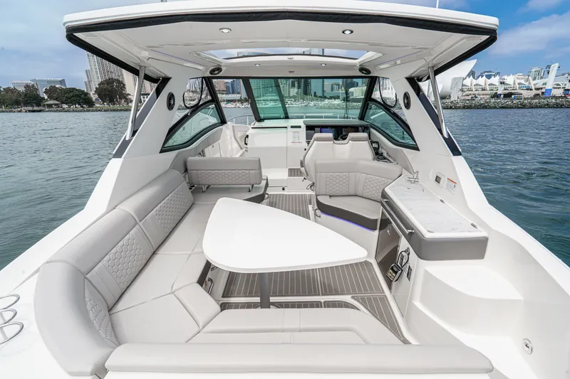 Slide: The Image of The image shows the cockpit of a 2025 Sea Ray 320 Sundancer DA Coupe. It features sleek, modern interior with gray upholstery and a white table. The setting is a calm harbor with a cityscape in the background. - 2