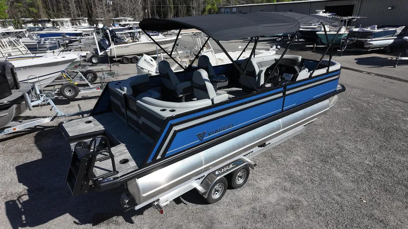 Slide: The Image of 2025 Viaggio Z24Q pontoon boat with blue accents on a trailer in a marina. - 1