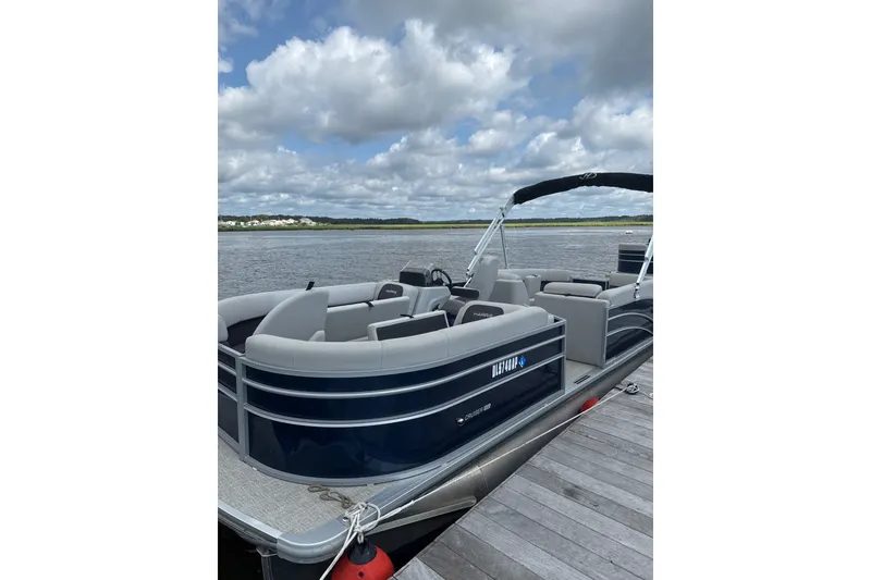 The Image of 2024 Harris Cruiser 230 pontoon boat docked on a scenic lake under a cloudy sky. - 1