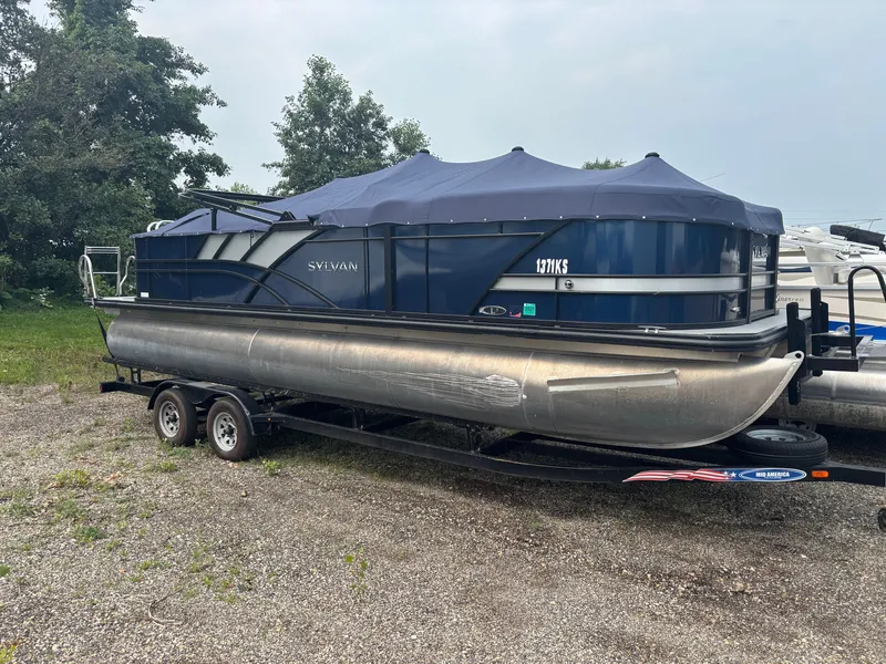 Slide: The Image of A 2021 Sylvan L3PF TRITOON on a trailer, with a blue cover, parked on a gravel lot. - 1