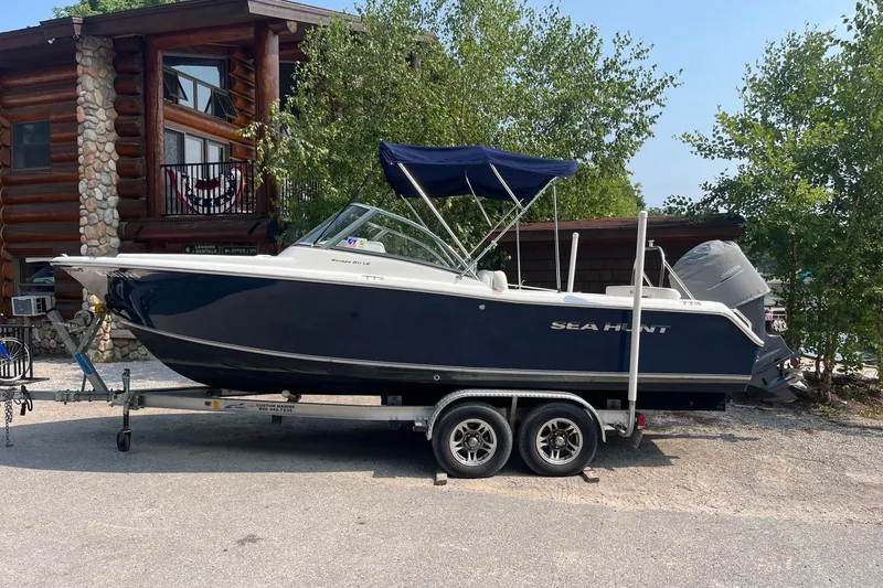 The Image of 2014 Sea Hunt Escape 211 LE boat on trailer, parked near a rustic log cabin. - 1