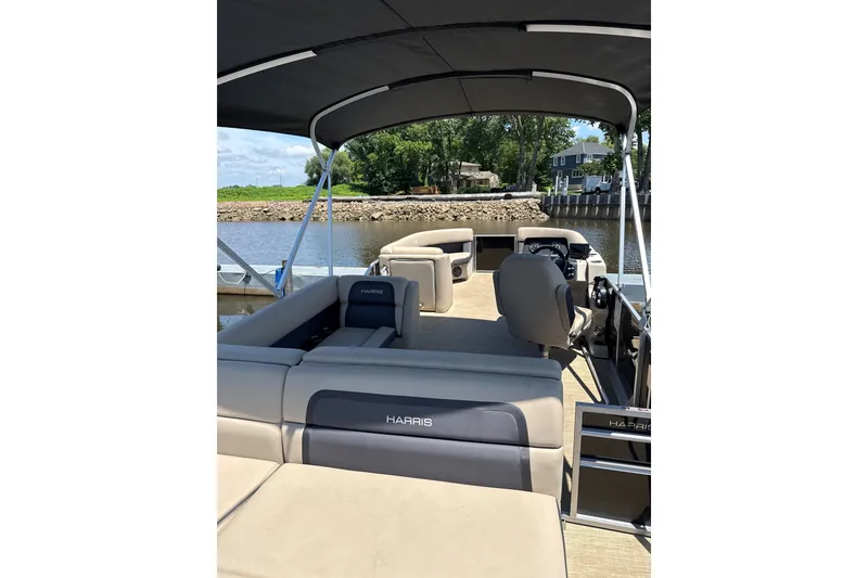 Slide: The Image of 2024 Harris Cruiser 230 pontoon boat interior with seating and helm, docked by a scenic river. - 7