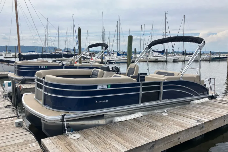 Slide: The Image of 2024 Harris Cruiser 230 pontoon boat docked at a marina with sailboats in the background. - 2