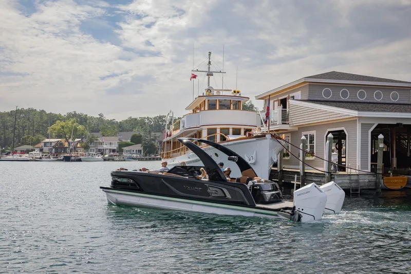 The Image of 2026 Tahoe Pontoon Grand Tahoe Elite Windshield 25 cruising near a docked boat and boathouse. - 3