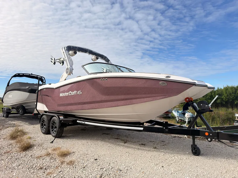 Slide: The Image of A 2027 MasterCraft NXT22 boat on a trailer on a gravel path under a blue sky. - 2