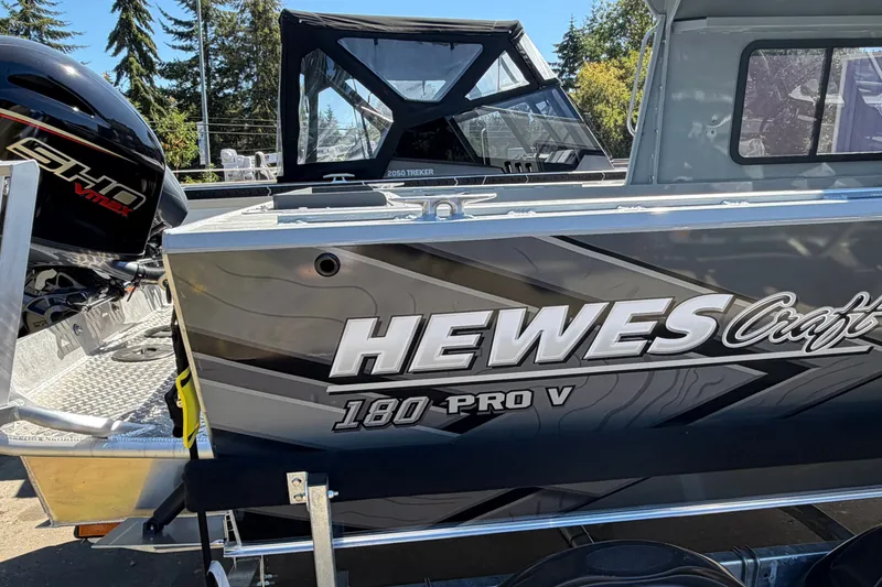 Slide: The Image of A 2026 Hewescraft 180 ProV Hard Top boat with an outboard motor, parked outdoors. - 2
