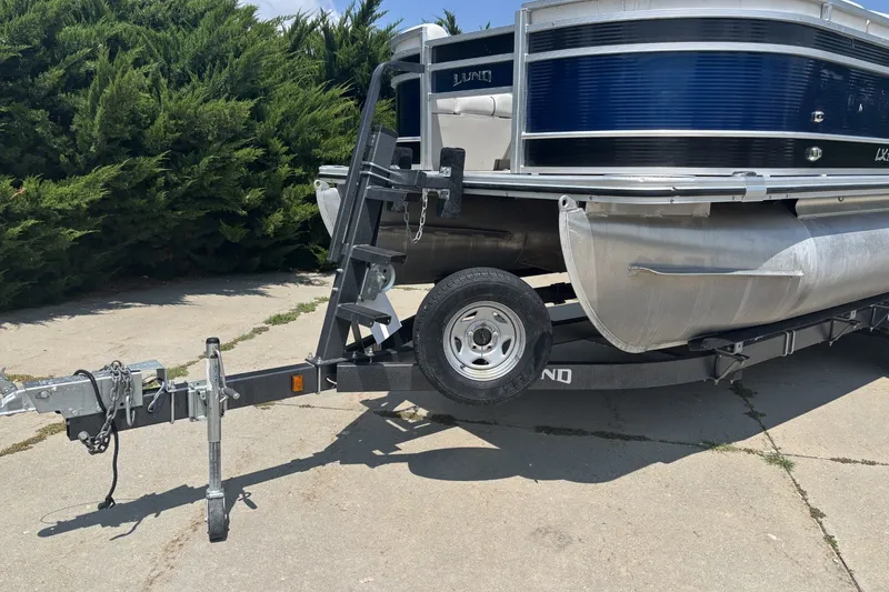 Slide: The Image of 2020 Lund LX220 Pontoon on trailer, parked on concrete, surrounded by greenery. - 16