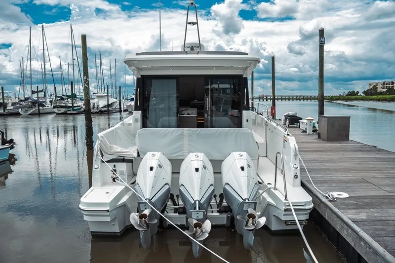 Slide: The Image of 2025 Jeanneau NC 1295 Coupe docked, featuring triple outboard engines, under a cloudy sky. - 10