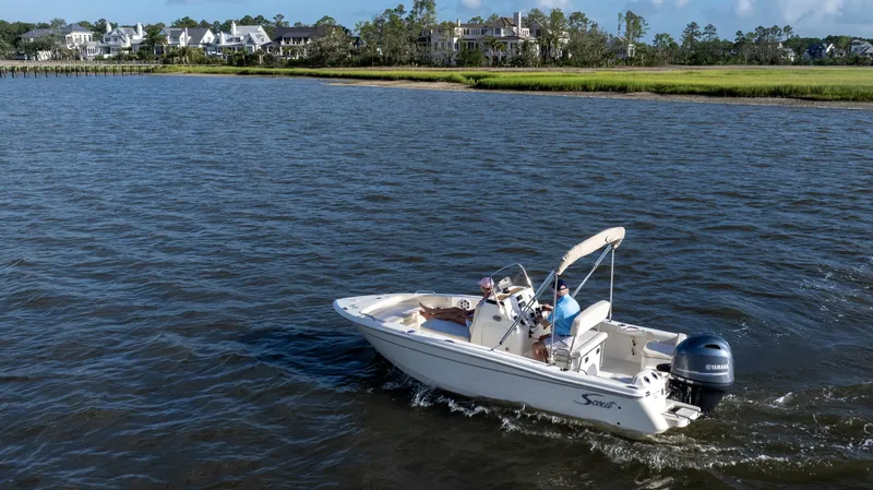 Slide: The Image of Scout 188 Sportfish 2027 on calm water, with two people aboard, near a shoreline with houses. - 38