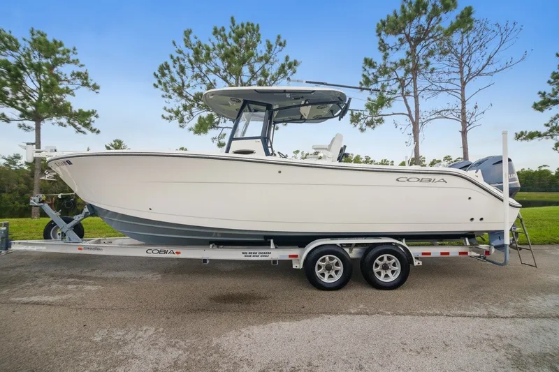 The Image of 2021 Cobia 280 Center Console boat on trailer, parked outdoors with trees in background. - 0
