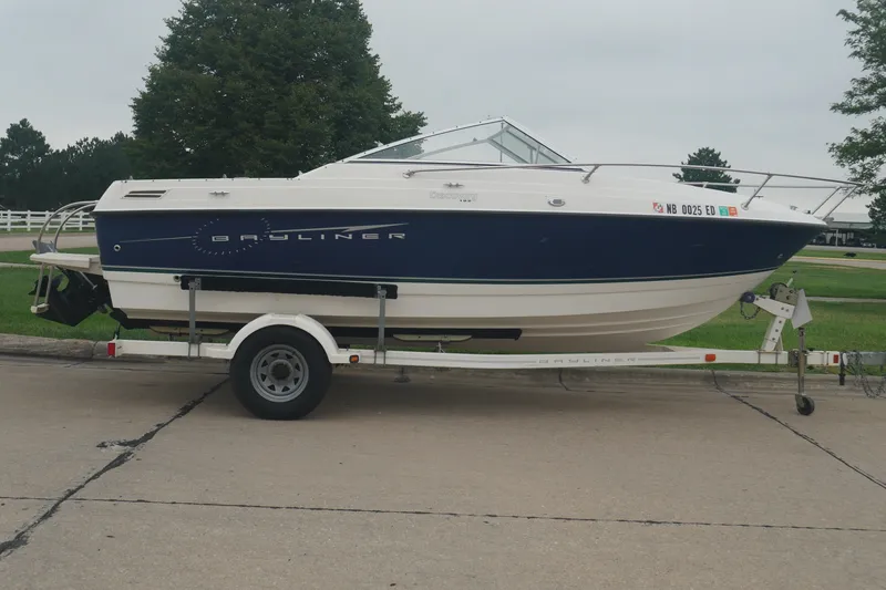 The Image of 2008 Bayliner 192 Discovery boat on trailer, parked on a paved surface with greenery background. - 0