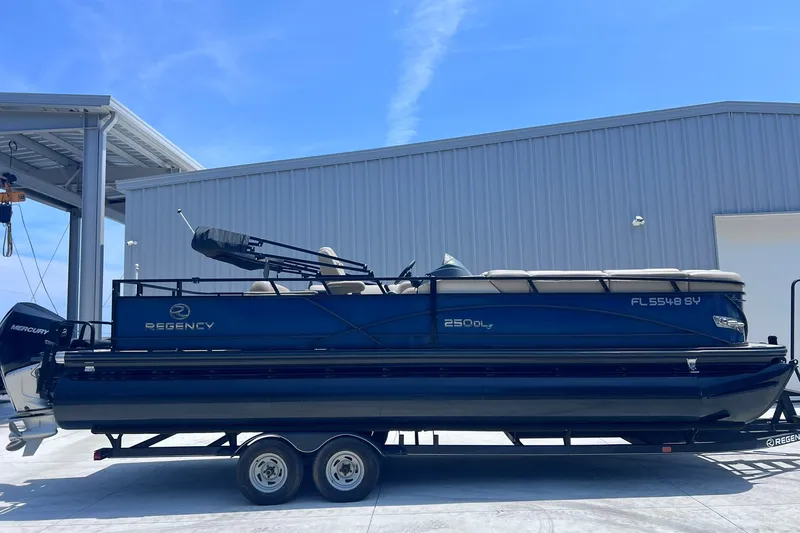 The Image of 2020 Regency 250 DL3 pontoon boat on trailer, parked near a metal building. - 1