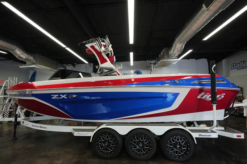 The Image of 2021 Tigé 25ZX boat in showroom, red and blue design on trailer. - 1