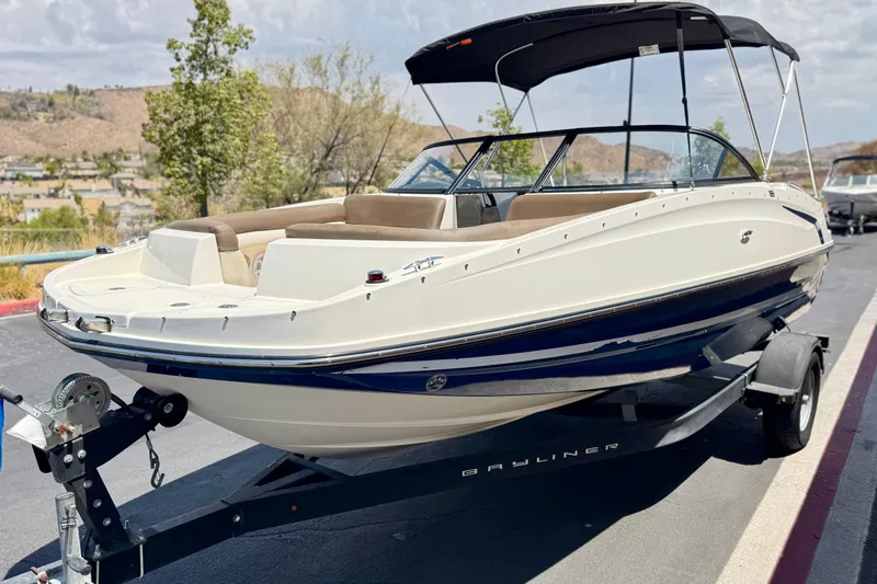 Slide: The Image of 2019 Bayliner 195 Deck Boat on trailer, parked outdoors, with bimini top and sleek design. - 3