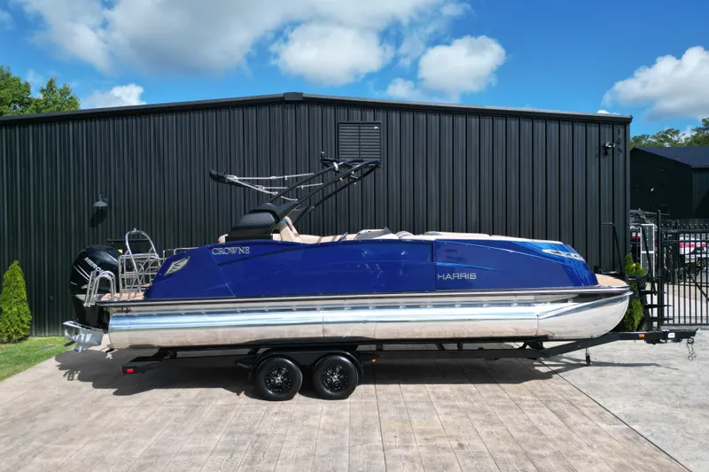 The Image of 2019 Harris Crowne SL 250 pontoon boat on trailer, parked by black building. - 0