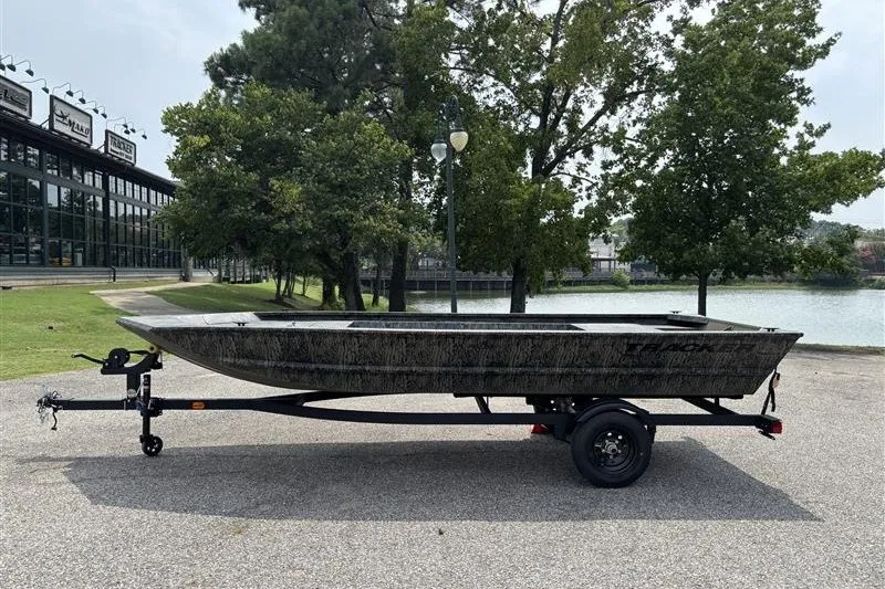 The Image of 2027 Tracker Grizzly 1648 Utility boat on trailer, parked near a lake with trees. - 0