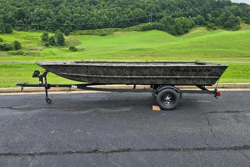The Image of 2027 Tracker Grizzly 1648 Utility boat on trailer, parked on asphalt with green hills background. - 0