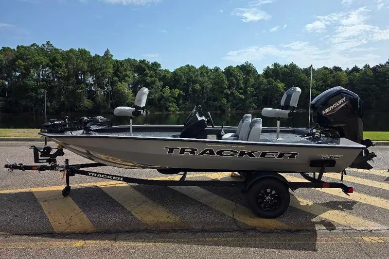 The Image of 2027 Tracker Pro Team 175 boat on trailer, parked near a lake with trees in background. - 0