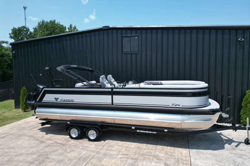 Slide: The Image of 2027 Viaggio Lago X-S 24 pontoon boat on trailer, parked by a black building. - 22