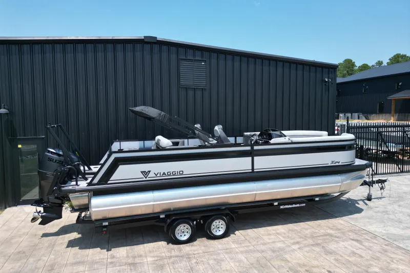 Slide: The Image of 2027 Viaggio Lago X-S 24 pontoon boat on trailer, parked by a black building. - 23