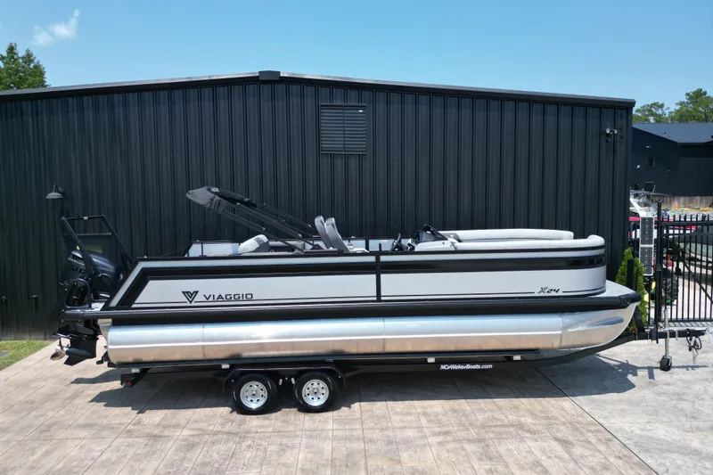 Slide: The Image of 2027 Viaggio Lago X-S 24 pontoon boat on trailer, parked by black building. - 24