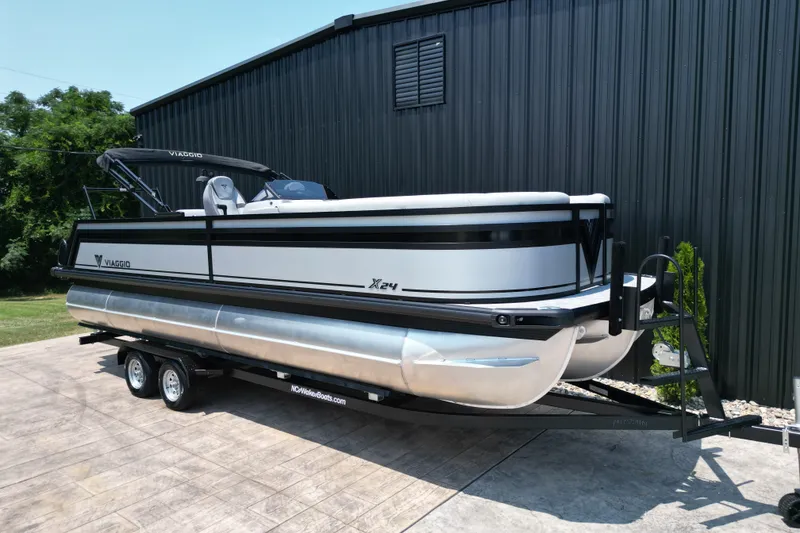 Slide: The Image of 2027 Viaggio Lago X-S 24 pontoon boat on trailer, parked by a black building. - 19